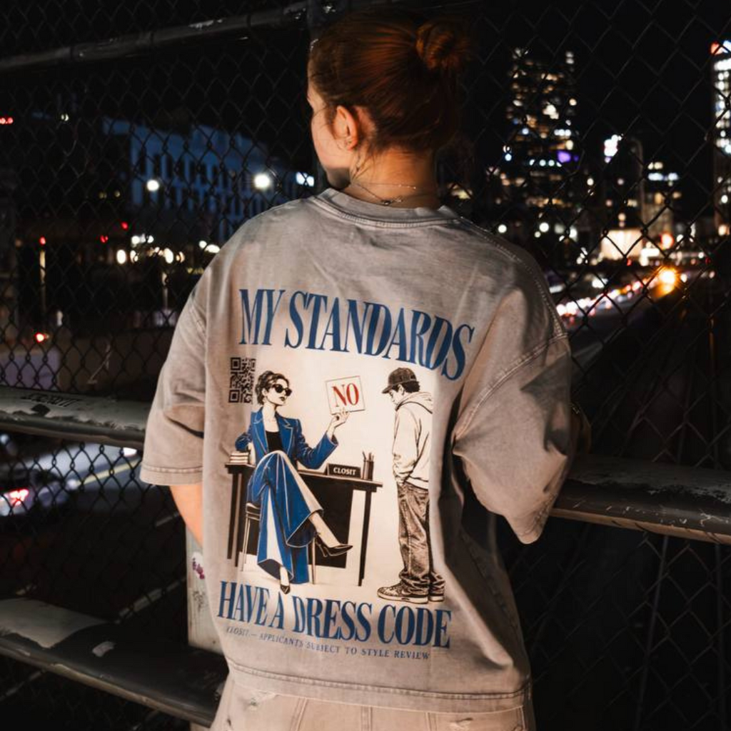 Back view of a woman wearing the light gray My Standards Have a Dress Code graphic tee at night.